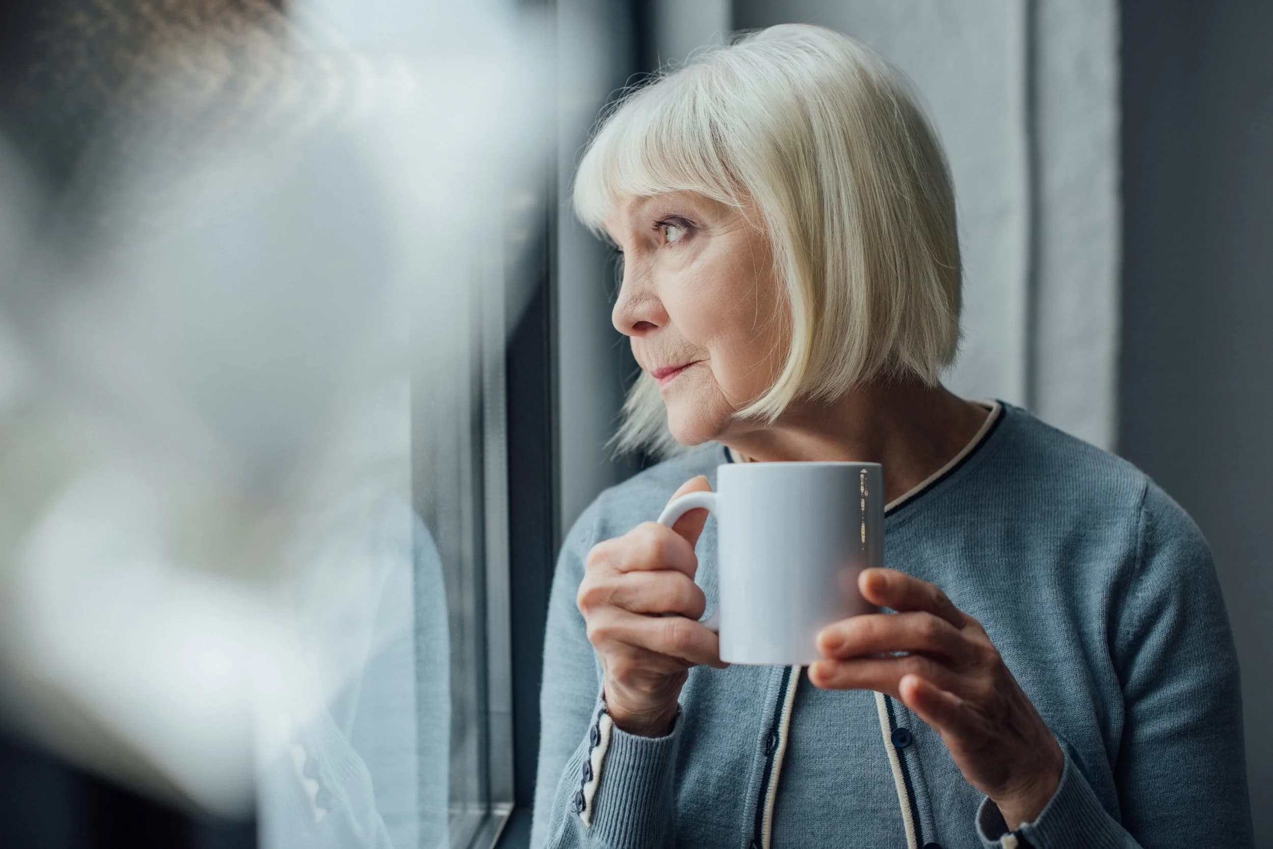 Ältere Frau mit einer Tasse schaut nachdenklich aus dem Fenster