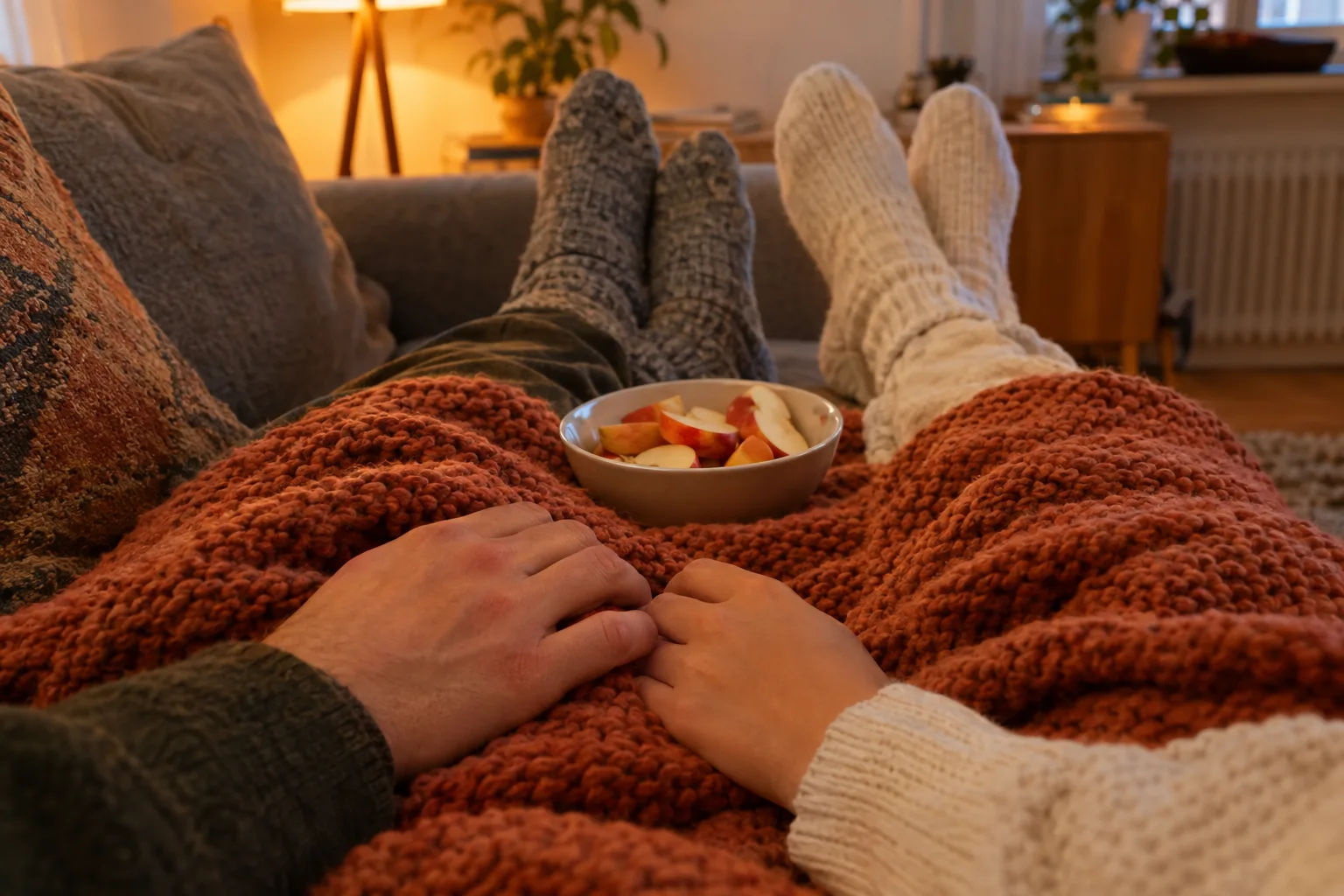 Eine rostrote Strickdecke bedeckt die Beine zweier Personen auf dem Sofa, ihre Hände liegen nebeneinander auf der Decke.